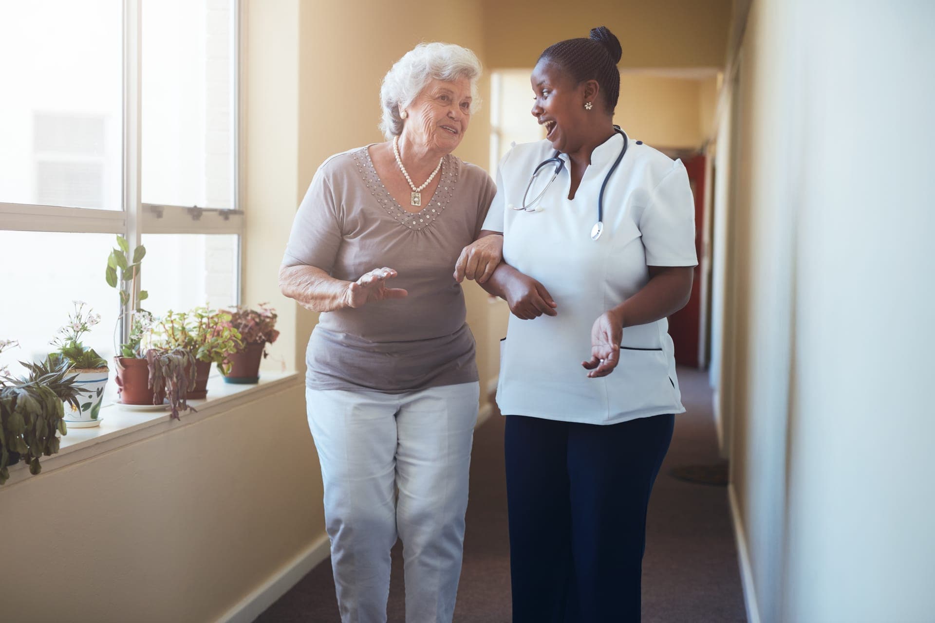 Caregiver helping an elderly woman walk down a hallway, both talking